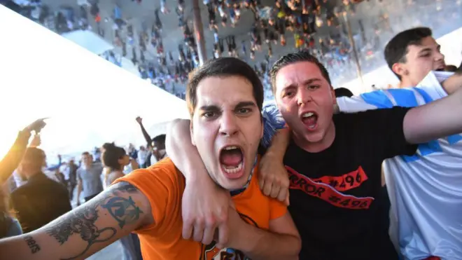 مشجعان يغمران بعضهما في مباراة لكرة القدم، ويصرخان، على اليمين شاب بقميص أسود وعلى اليسار شاب بقميص برتقالي مع وشم لفريق ماريسيليا الفرنسي على ذراعه