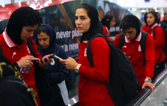 İran kadın milli futbol takımının oyuncuları endişeli şekilde telefonlarına bakıyor. Hepsi kırmızı giymiş ve havalimanı yürüyen merdivenindeler.