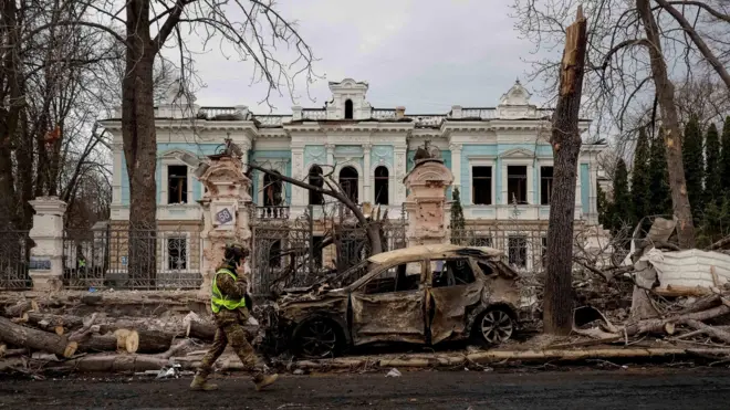 Сумы. На заднем плане старое здание с выбитыми стеклами, на переднем сгоревшая машина и обломки деревьев, мимо проходит человек в военной форме.