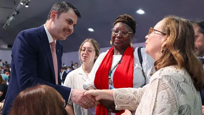 Türk bakan takım elbiseli karşısındaki kadının gözlerinin içine bakıyor, yanında Afrika kökenli gözlüklü bir kadın var. 