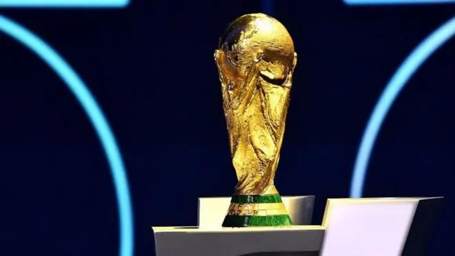 サッカー男子W杯のトロフィー。台座は三角錐の下半分の形で、緑と金色に塗り分けられ、金色部分に「FIFA WORLD CUP」の文字が見える。トロフィー部分は金色で、4人の人物が大きなサッカーボールを掲げているような彫刻になっている