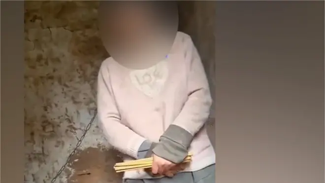 A screenshot of the video shows the woman with a chain around her neck standing in the hut
