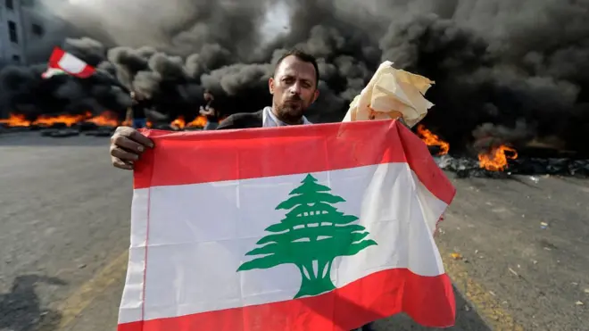 متظاهر يحمل علم لبنان خلفه حاجز من الإطارات المطاطية المشتعلة على الطريق الرابطة بين بيروت ومدينة طرابلس شمالا يوم الجمعة الثامن عشر من أكتوبر تشرين الأول