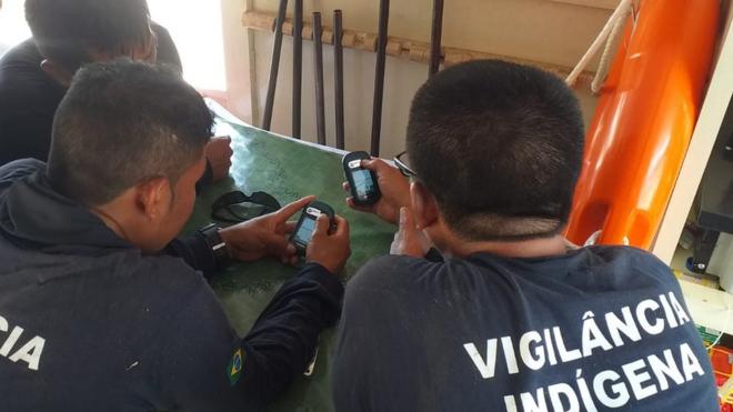 Fotografia colorida mostra homens indígenas de costas olhando para um pequeno aparelho eletrônico