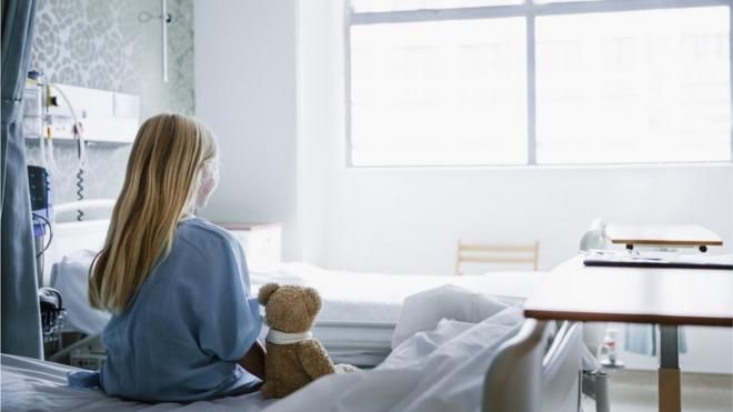 فتاة صغيرة تجلس على سرير المستشفى وبجانبها دمية دب