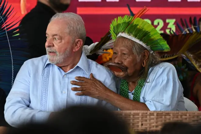 Lula ao lado de cacique Raoni durante evento do governo. Raoni coloca a mão no peito de Lula, que está olhando para o lado