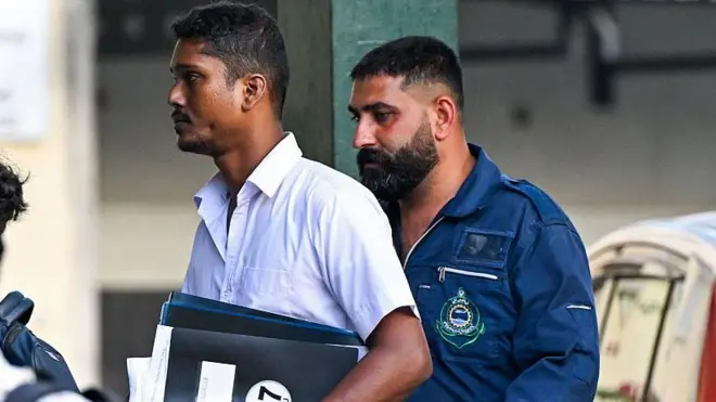 Bir İranlı denizci Sri Lankalı yetkilinin hemen arkasında yürüyor. Sağlıklı görünüyor.