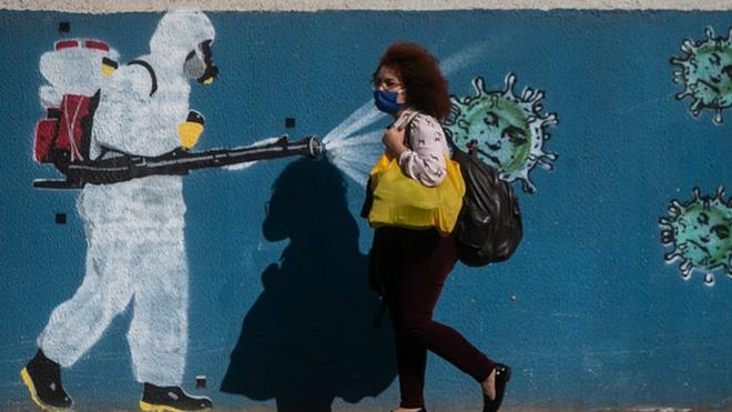 A woman wearing a mask walks past a wall with a graffiti depicting a cleaner in protective gear spraying viruses with the face of President Jair Bolsonaro