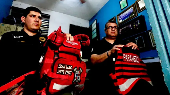 Dos paramédicos, Julio César Vega y Héctor Torres, están sentados en una pequeña habitación. Llevan su equipo sobre las rodillas. Ambos visten uniformes negros y Torres lleva gafas.