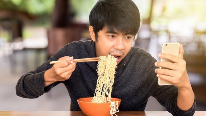 Hombre comiendo noodles y revisando su celular