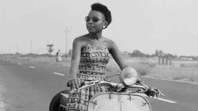 A woman sitting on a moped by the roadside poses for the camera.