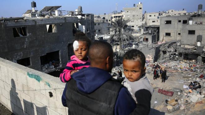 A man holds two children as he looks at the scene of a reported Israeli air strike in Nuseirat refugee camp, in the central Gaza Strip (18 December 2023)
