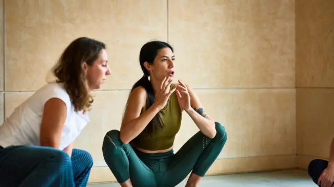 İki kadın yoga stüdyosuna benzer bir odada çömelme hareketi yapıyor. 
