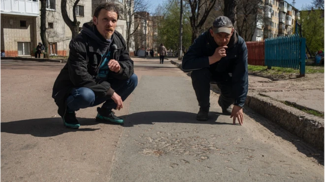 Two eyewitnesses from the bread queue attack kneel beside a classic cluster impact mark.