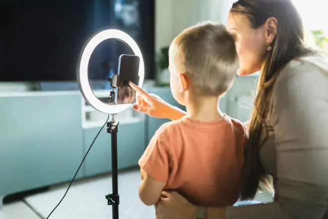 Mãe ao lado de menino; eles estão de costas, em frente a celular e ring light