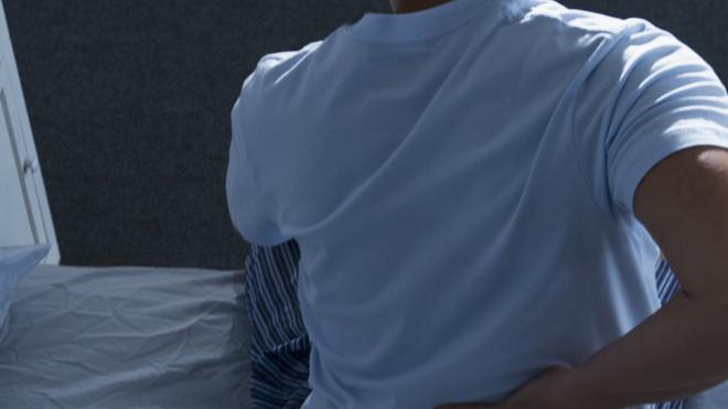 Pessoa sentada na cama tocando a lombar com a mão