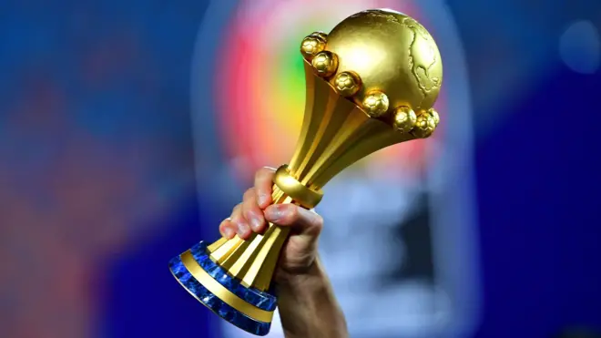 The Africa Cup of Nations trophy is held aloft by a man's right hand in front of a background which is out of focus