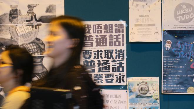 香港浸会大学逸夫校园内一群女学生走过贴上了反对普通话毕业门槛豁免试标语的学生会民主墙（BBC中文网图片22/1/2018）