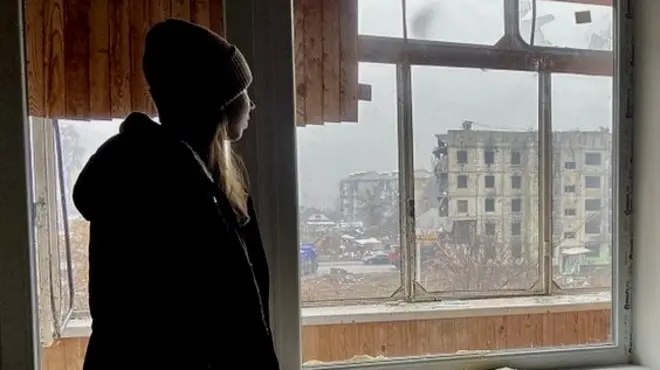 Анастасія у квартирі батьків, де родина не живе після того, як вибухова хвиля пошкодила будинок. З балкону видно розбомблений будинок, від вибуху в якому постраждав дім Анастасії.