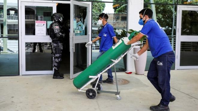 Dois homens de máscara carregam carrinho com tanque de oxigênio, em frente a porta de hospital