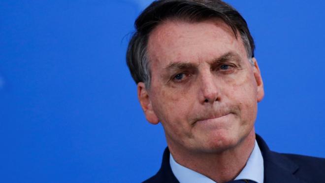Bolsonaro em frente a painel azul de evento cerrando os lábios
