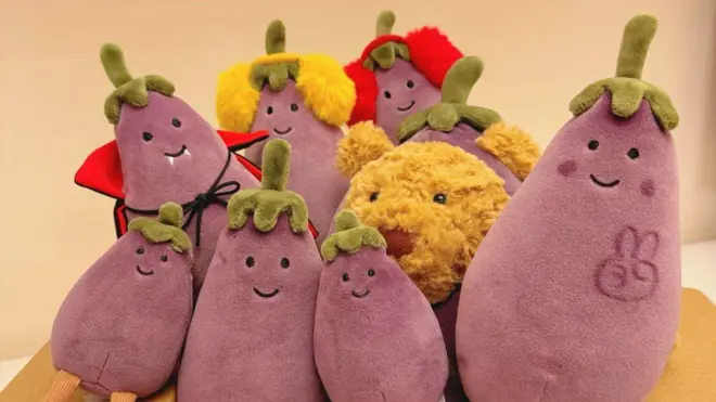 Eight Jellycat aubergine plushies of different sizes surround a teddy bear at the centre. A peace sign is drawn on the rightmost aubergine