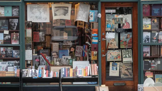 通り沿いの書店のウィンドウや入口にポスターが貼られ、ディスプレイにさまざまな本が並べられている。ウィンドウ越しに店内の書棚も見える。店の外にも低い棚が置かれ、本が並べられている