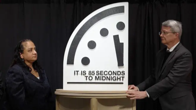 Two scientists are holding a clock that says "85 seconds to midnight."