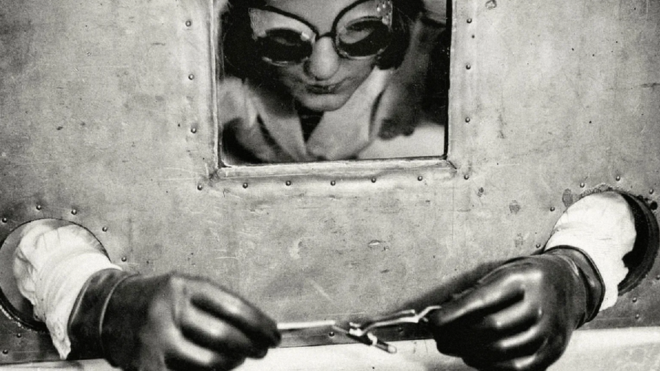 Mulher manipulando material radioativo usando luvas e uma barreira para proteção, em foto preto e branco