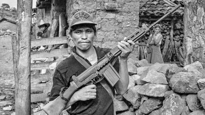 Un miliciano rebelde alza su rifle en un gesto de desafío frente a su casa en el departamento de Chalatenango, ocupado por los rebeldes, El Salvador, 1984. Foto por Scott Wallace.