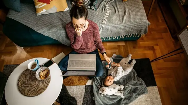 Una mujer frente una computadora portátil trabajando y atendiendo a su bebé a la vez