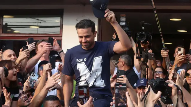 Foto colorida mostra o empresário Pablo Marçal em meio a uma multidão de pessoas que tiram fotos dele com seus celulares. Ele uma uma camiseta azul com a letra M e levanta um boné azul em um gesto de cumprimento