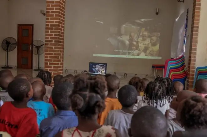 Les pensionnaires de l'orphelinat, de dos, regardent un film projeté sur grand écran dans un hall.