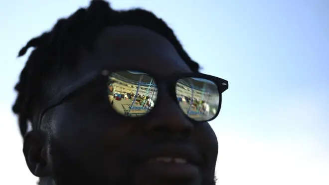 A man stands in the shade with bright reflections on his sunglasses.