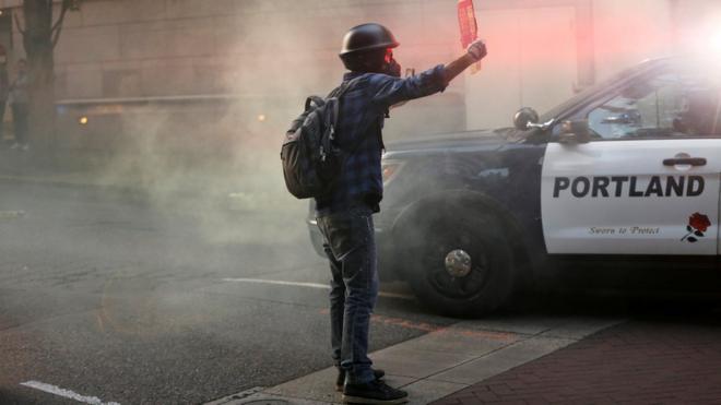 Jovem protesta em Portland e segura cartaz em frente a carro de policia