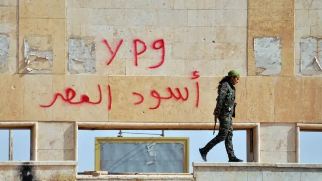 Duvarda kırmızı grafiti, YPG yazıyor, önünde silahlı bir kadın militan yürüyor