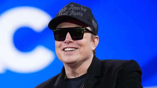 Tech multi-billionaire Elon Musk dey tok for di Conservative Political Action Conference (CPAC) for February 2025. E wear all black, plus one Make America Great Again baseball cap, coat, t-shirt and sunglasses. Di background na bright blue wit on blue large white letter C ova Musk right shoulder.