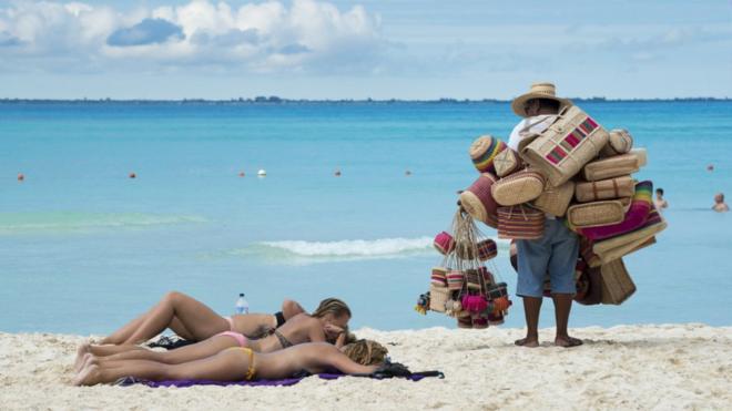 Mujeres tomando sol en la playa mientras un vendedor las mira en México.