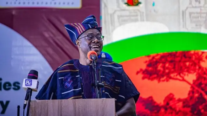 Gomina Seyi Makinde wọ aṣọ ofi, gbohungbohun wa niwaju rẹ, o n wo ẹgbẹ to si n rẹrin-in