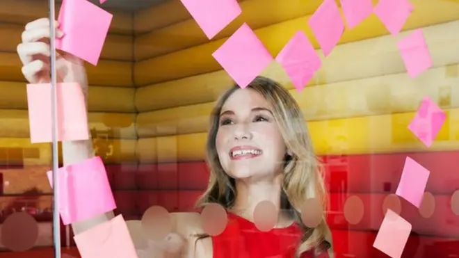 Una mujer haciendo un corazón de post its en un vidrio de una oficina