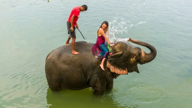 An elephant is splashing water on a tourist woman in the Rapti River in Chitwan National Park.