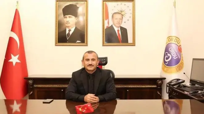 Siyah takım elbiseli bir adam makam odasında oturuyor ve yanında Türk bayrağı var.