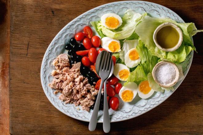 Salade française nicoise composée de thon en conserve, d'olives et d'œufs, servie avec une vinaigrette à l'huile d'olive et du sel de mer sur une assiette en céramique sur fond de bois.
