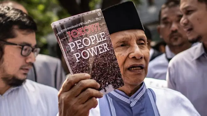 Ketua Dewan Kehormatan Partai Amanat Nasional (PAN) Amien Rais (tengah) menunjukkan buku berjudul Jokowi People Power saat jeda pemeriksaan untuk Shalat Jumat di Direktorat Reserse Kriminal Umum (Ditreskrimum) Polda Metro Jaya, Jakarta, Jumat (24/05).