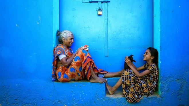 Пожилая женщина и девочка сидят в нише стены с котенком и курицей в руках