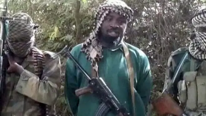 Abubukar Shekau stand between two militants wey hold machine gun.