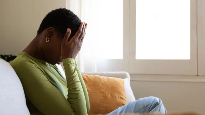 Young Black woman dey sit down for sofa, as e dey feel down and sad, come cover face wit hand.