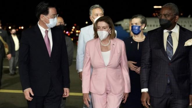 Pelosi caminha ao lado de outras pessoas com trajes formais; todos usam máscara e é noite
