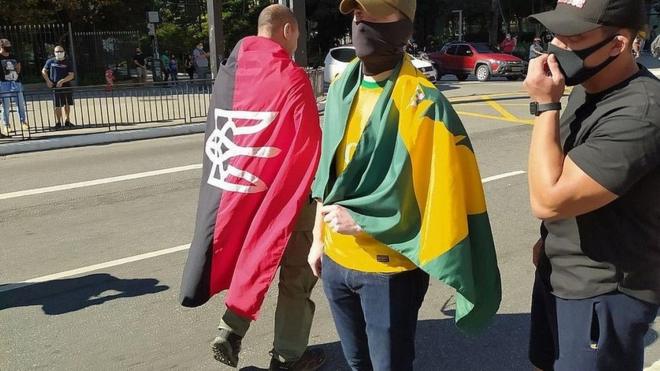 Manifestantes em protesto a favor de Bolsonaro escondem rosto e se enrolam em bandeiras; uma delas tem as corws vermelho e negra e o brasão nacional ucraniano