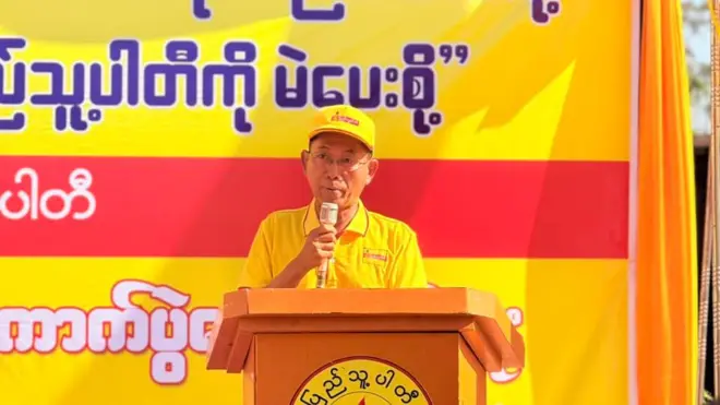 အဝါရောင်ဆိုင်းဘုတ်နောက်ခံနဲ့ စင်မြင့်ပေါ်မှာ ပါတီဦးထုပ်အဝါ၊ တီရှပ်အဝါဝတ်ပြီး မိန့်ခွန်းပြောနေတဲ့ ပြည်သူ့ပါတီဥက္ကဋ္ဌ ဦးကိုကိုကြီး 